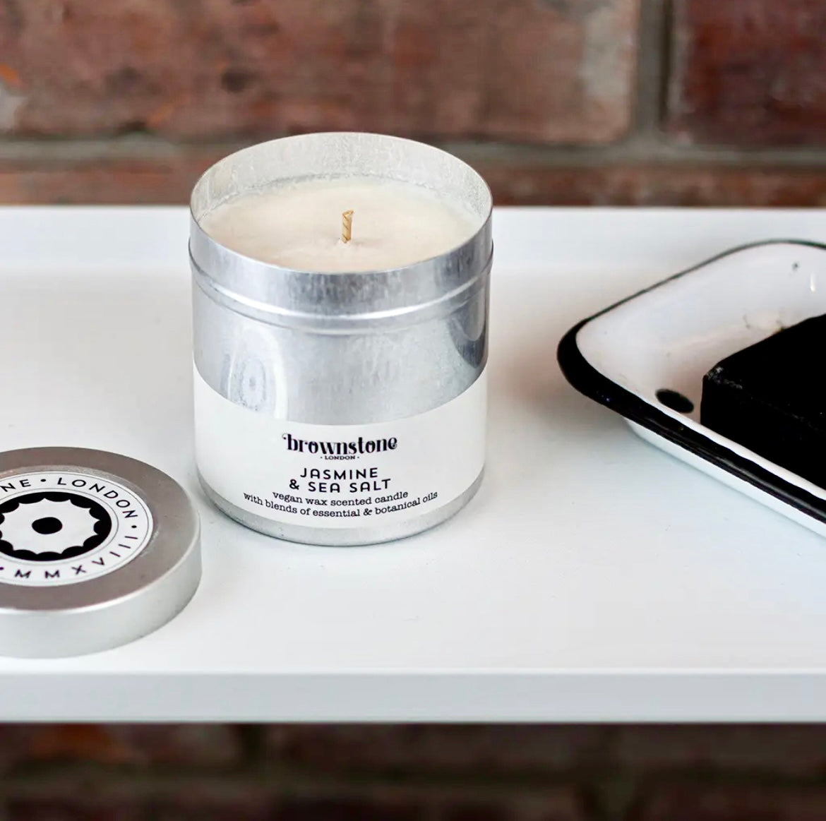 Candle in a metal container with a label on a white surface against a brick wall background