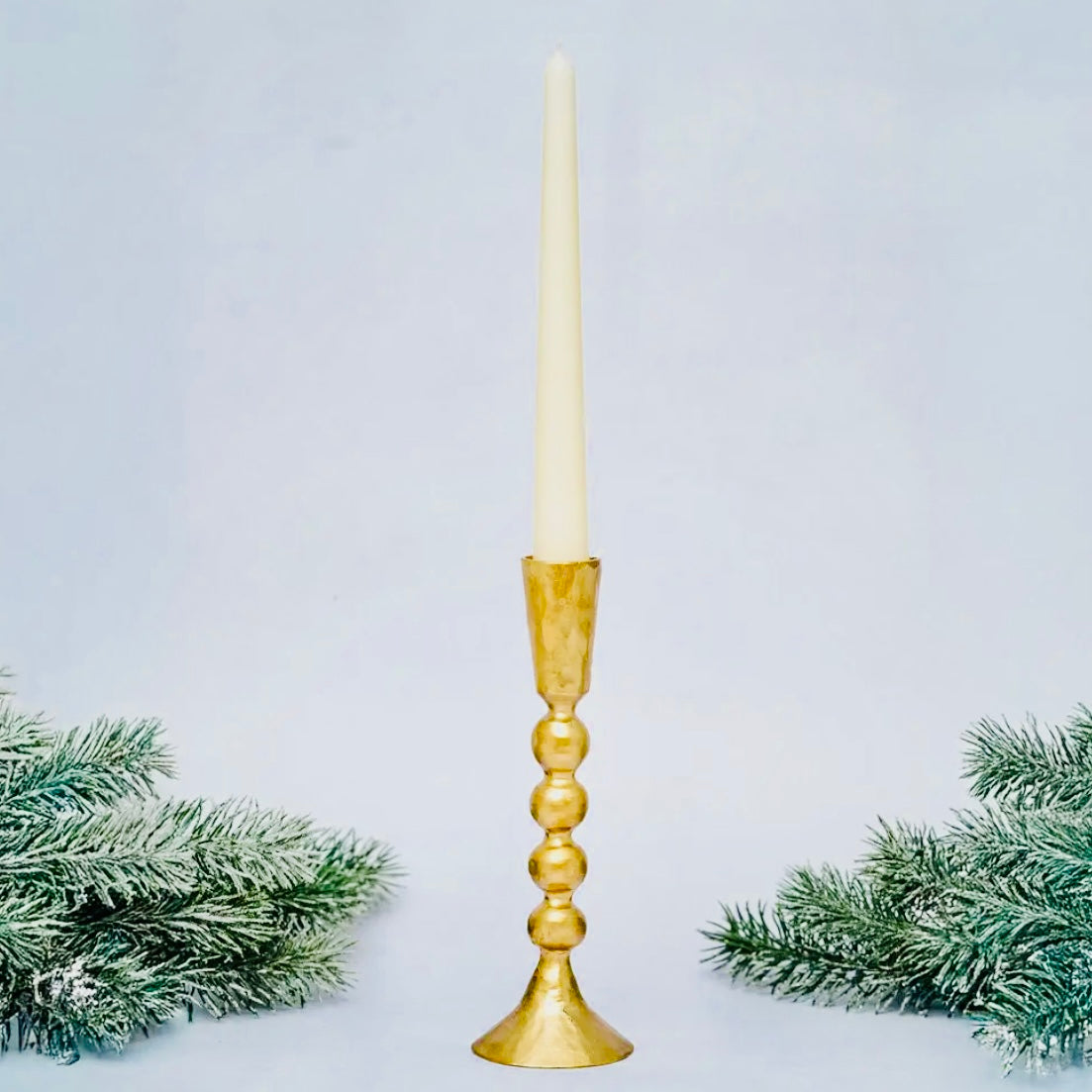 Gold candlestick with a white candle against a light blue background with decorative greenery.