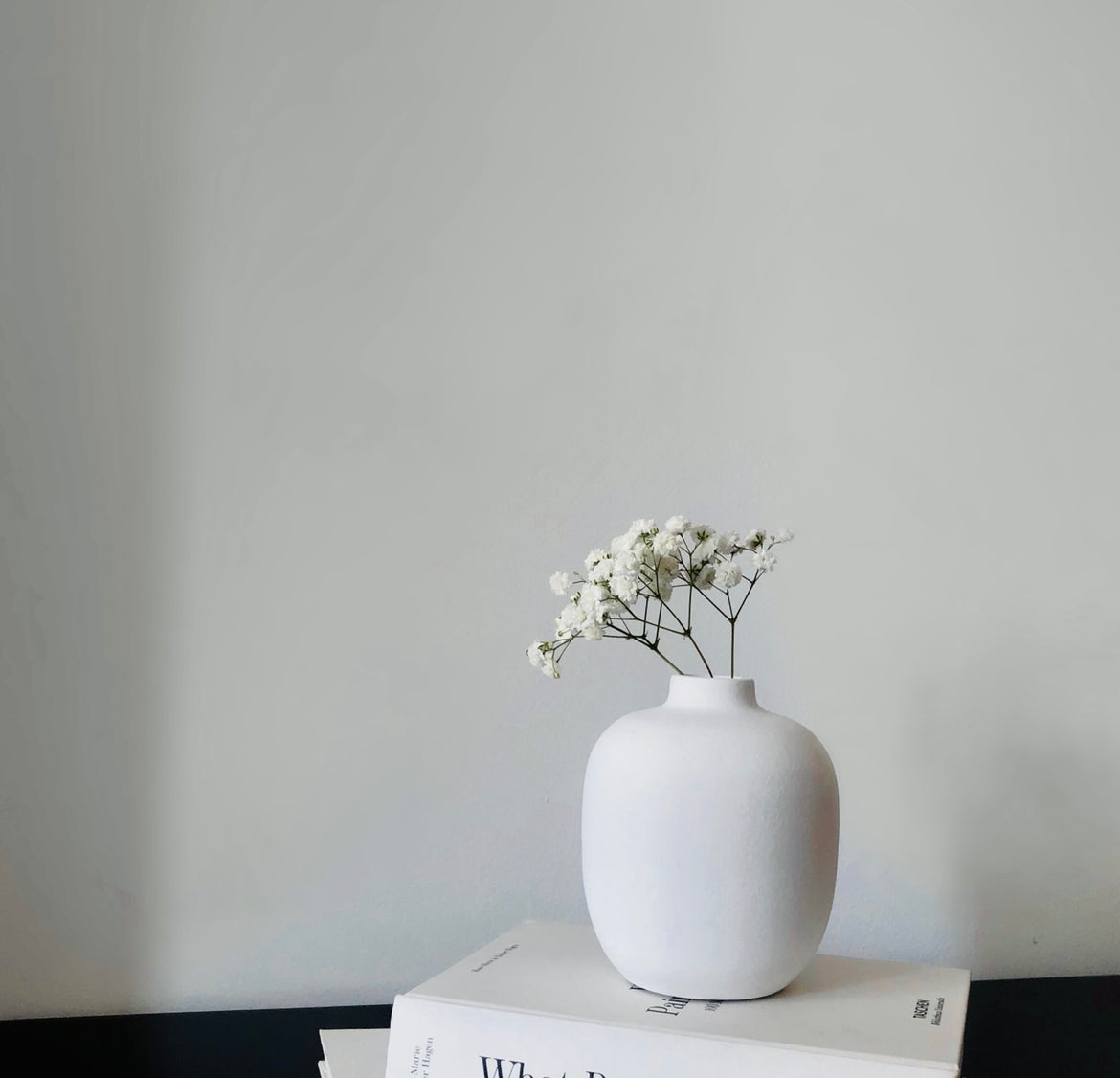 White vase with flowers on a stack of books against a plain wall