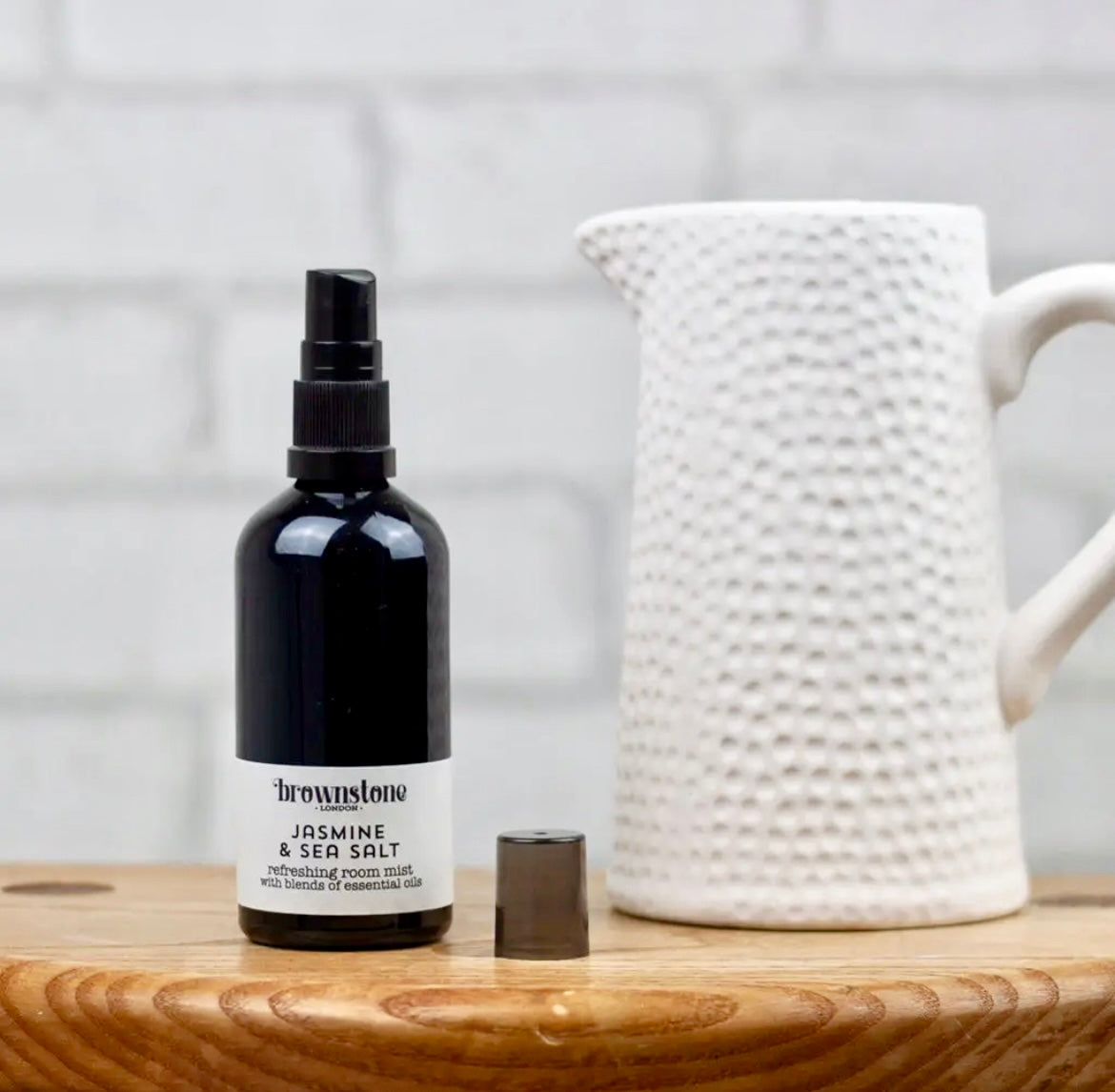 Black bottle of room mist with a white label on a wooden surface next to a white textured pitcher.