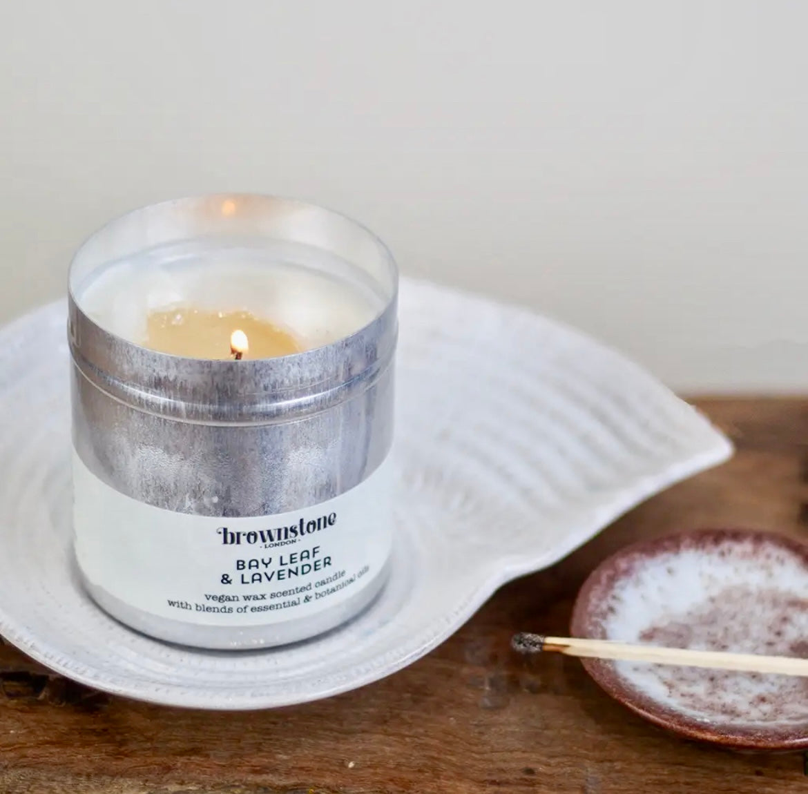 Candle in a metal container labeled 'Brownstone Bay Leaf & Lavender' on a white plate with a wooden surface.
