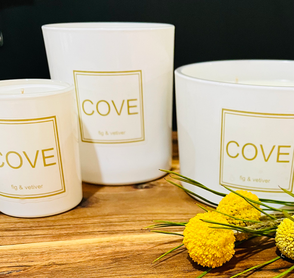 Three white candles labeled 'Cove' with gold text on a wooden surface, accompanied by yellow flowers.