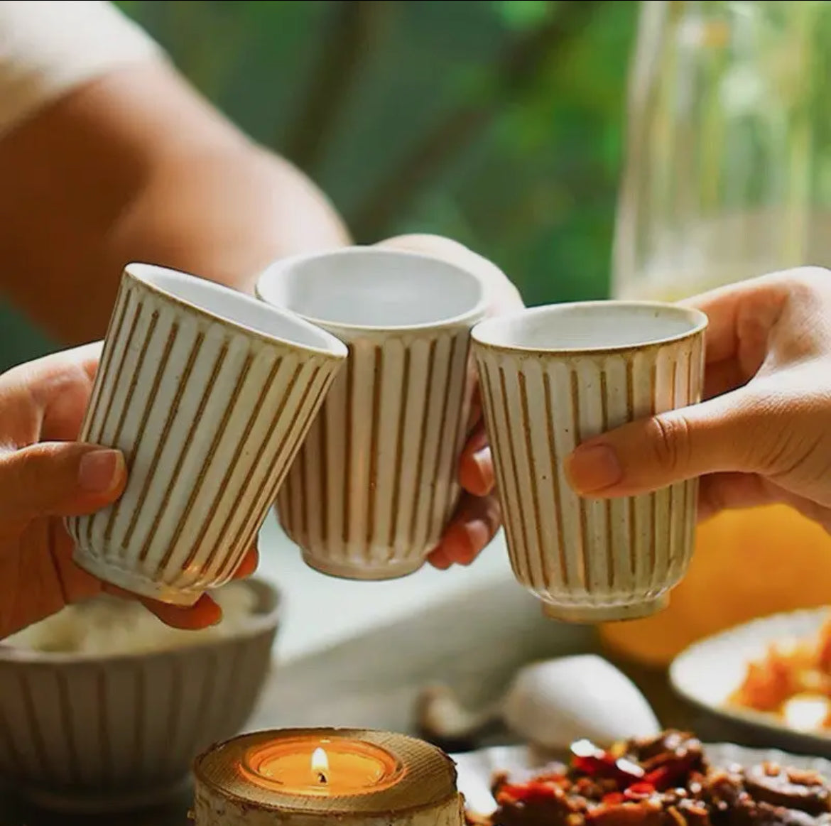 Three hands holding striped ceramic cups with a candle and food in the background