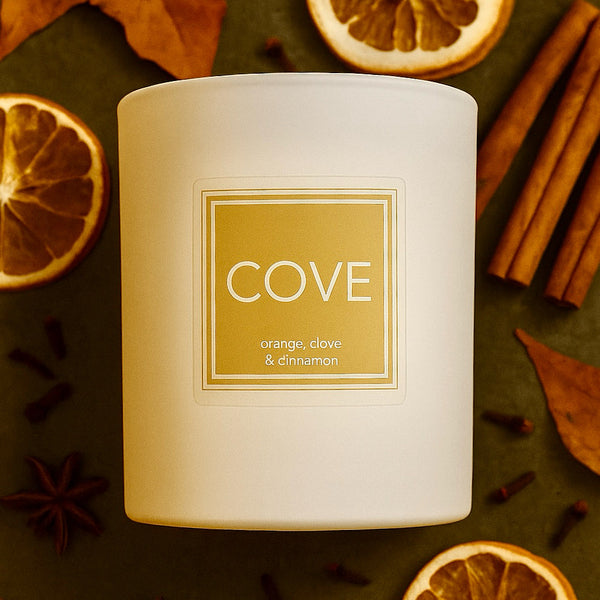Candle labeled 'Cove' with orange, clove, and cinnamon on a dark surface with dried oranges and cinnamon sticks.