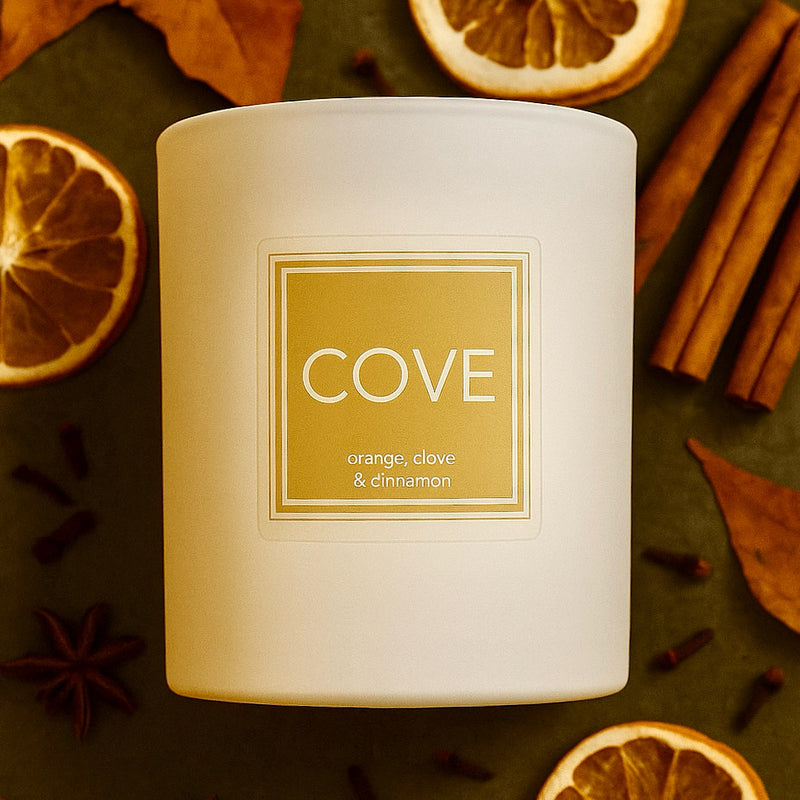 Candle labeled 'Cove' with orange, clove, and cinnamon on a dark surface with dried oranges and cinnamon sticks.