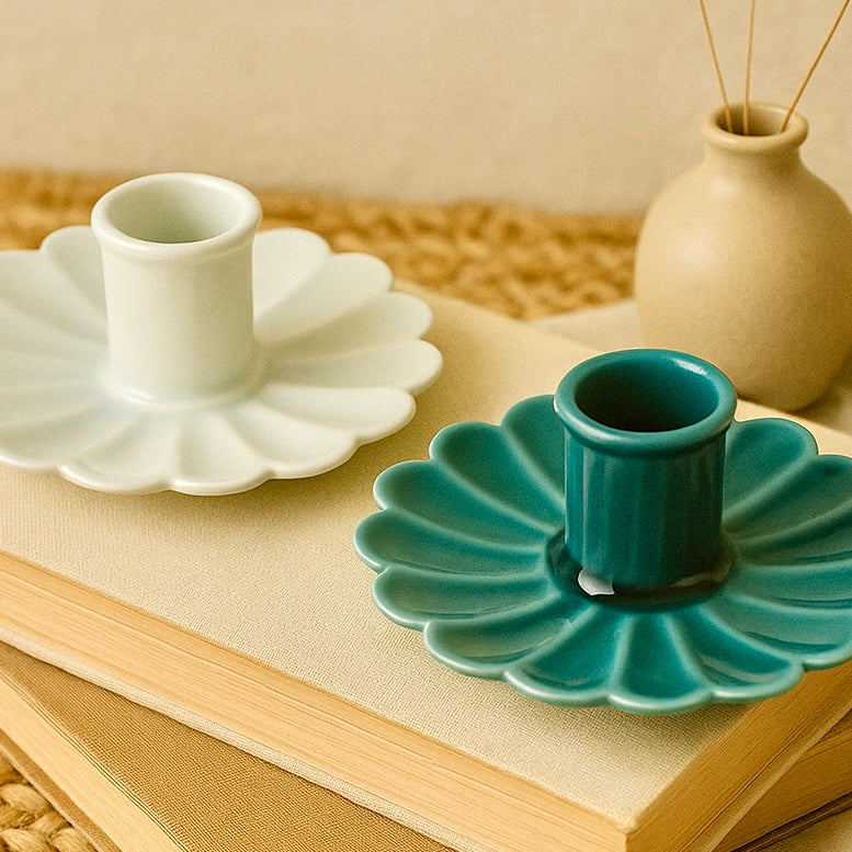 Two ceramic candle holders, one white and one teal, on a textured surface with a vase in the background.
