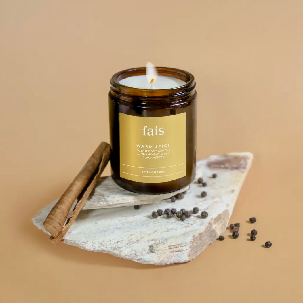 Fais Warm Spice Soy Candle in a brown jar with a label on a stone surface with cinnamon stick and peppercorns, against a beige background.