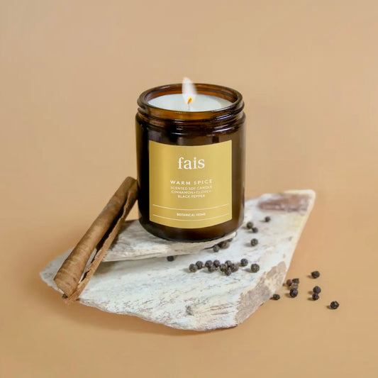 Fais Warm Spice Soy Candle in a brown jar with a label on a stone surface with cinnamon stick and peppercorns, against a beige background.