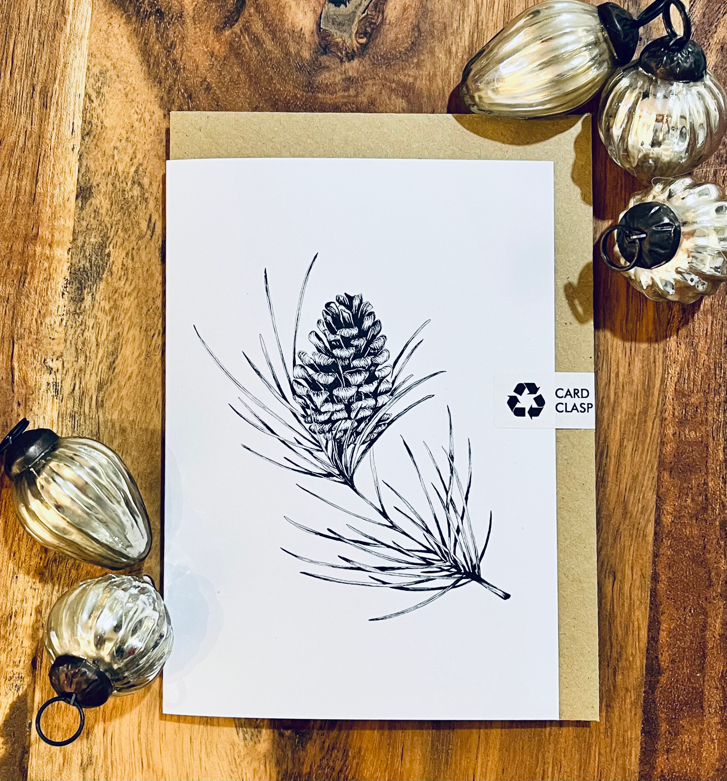 Greeting card with pine cone and branch illustration on a wooden surface with decorative pumpkins.