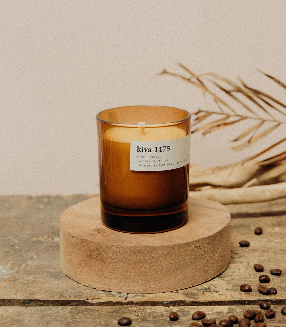 Amber glass candle labeled 'kiva 1475' on a wooden stand with coffee beans and branches in the background.