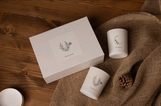 Two white candles with decorative labels on a wooden surface with burlap and pine cones.
