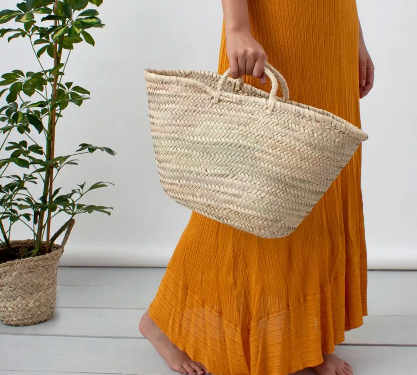 Person holding a woven basket wearing an orange dress next to a plant on a white floor.