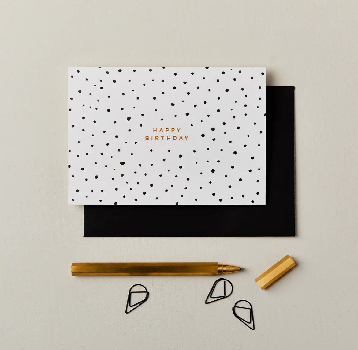 White card with black polka dots and 'Happy Birthday' text, black envelope, gold pen, and decorative items on a beige background.