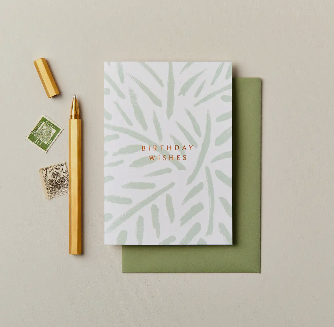 Greeting card with 'Birthday Wishes' text, green envelope, pen, and stamps on a beige background