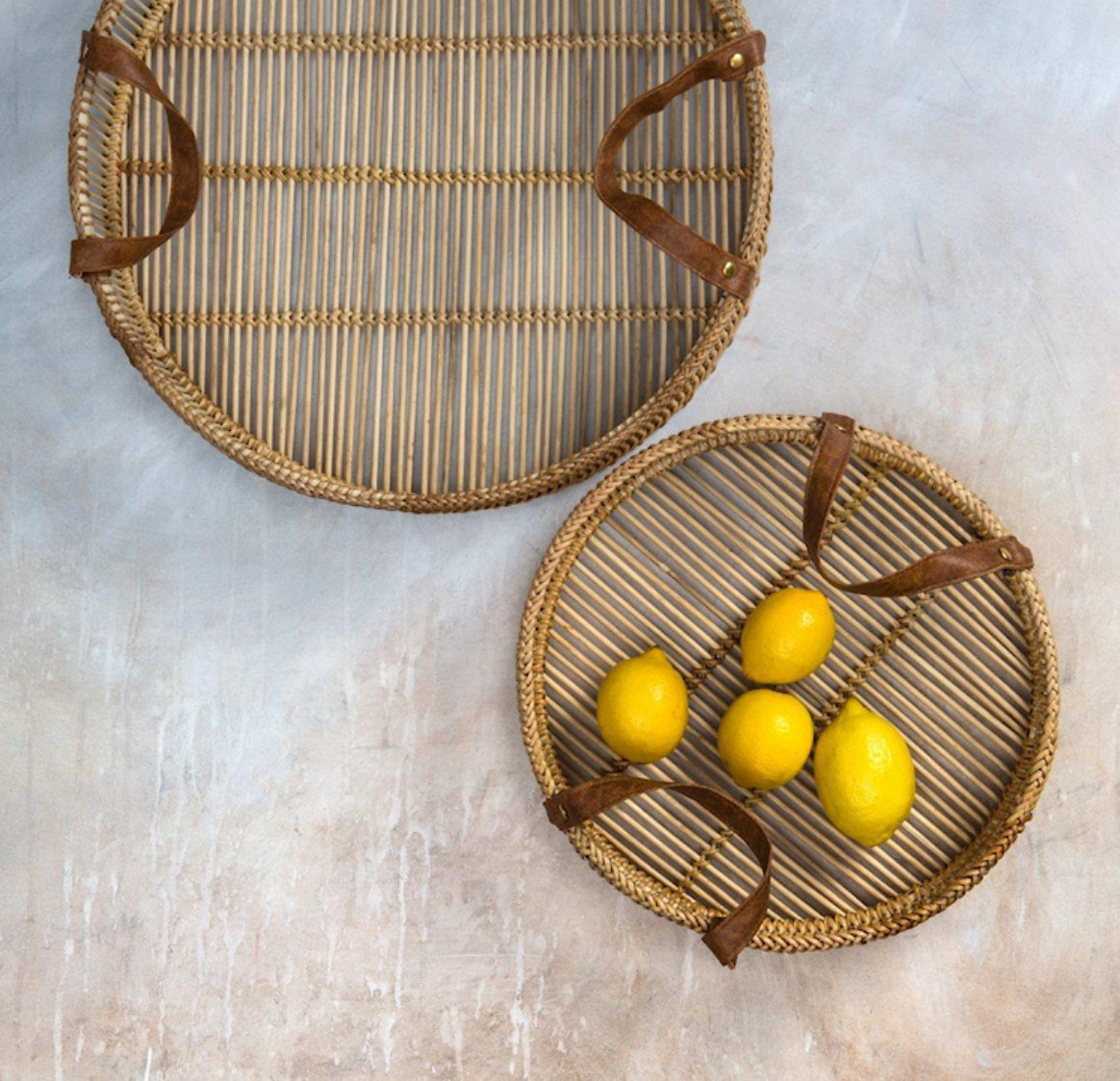 Two woven trays with brown handles on a light stone surface, one containing lemons.