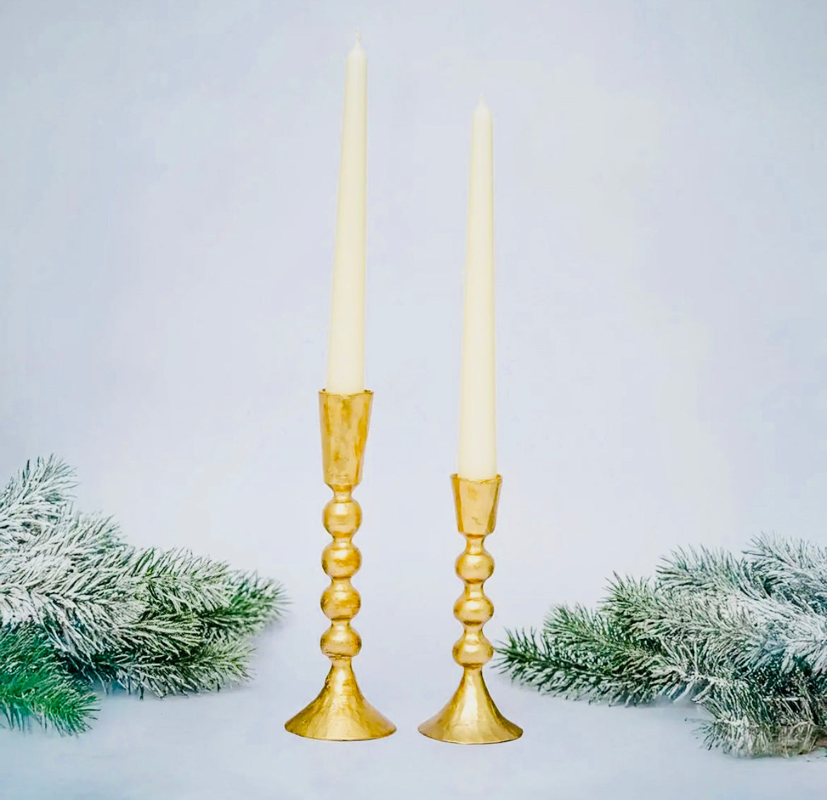 Two gold candlesticks with white candles on a light gray background with decorative greenery.